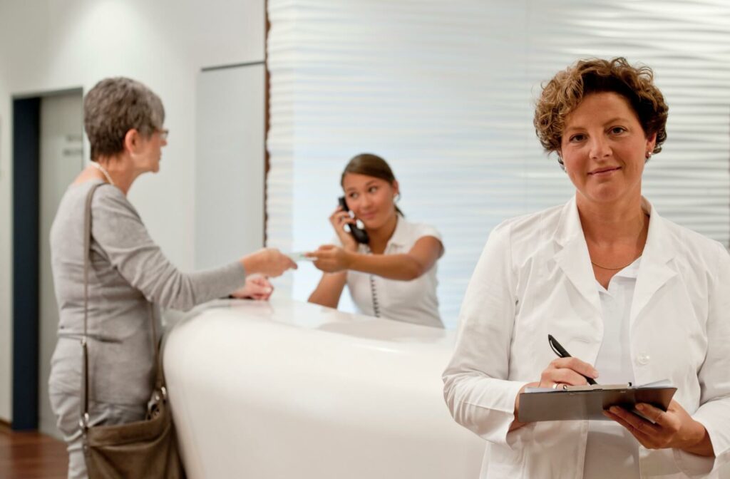 Senior woman handing over OHIP to receptionist at a clinic, with a eye doctor taking notes in the foreground.