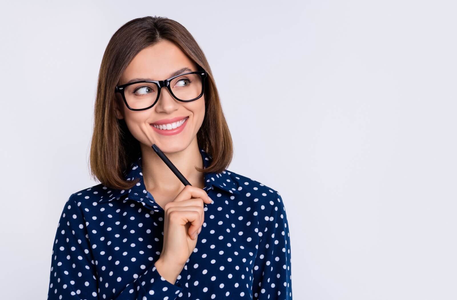 A smiling person wearing a polka-dotted shirt and bold black glasses taps their face thoughtfully with a pencil.