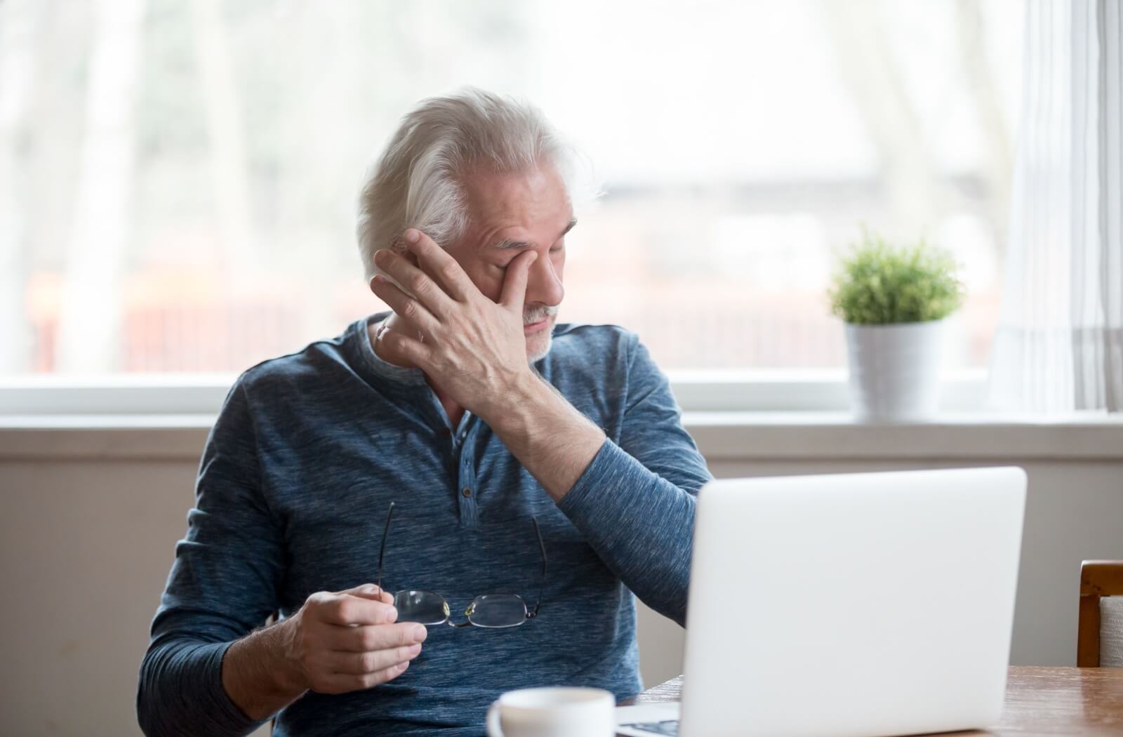 An older adult sitting at a desk rubbing their blurry eyes in frustration due to age-related macular degeneration (AMD).