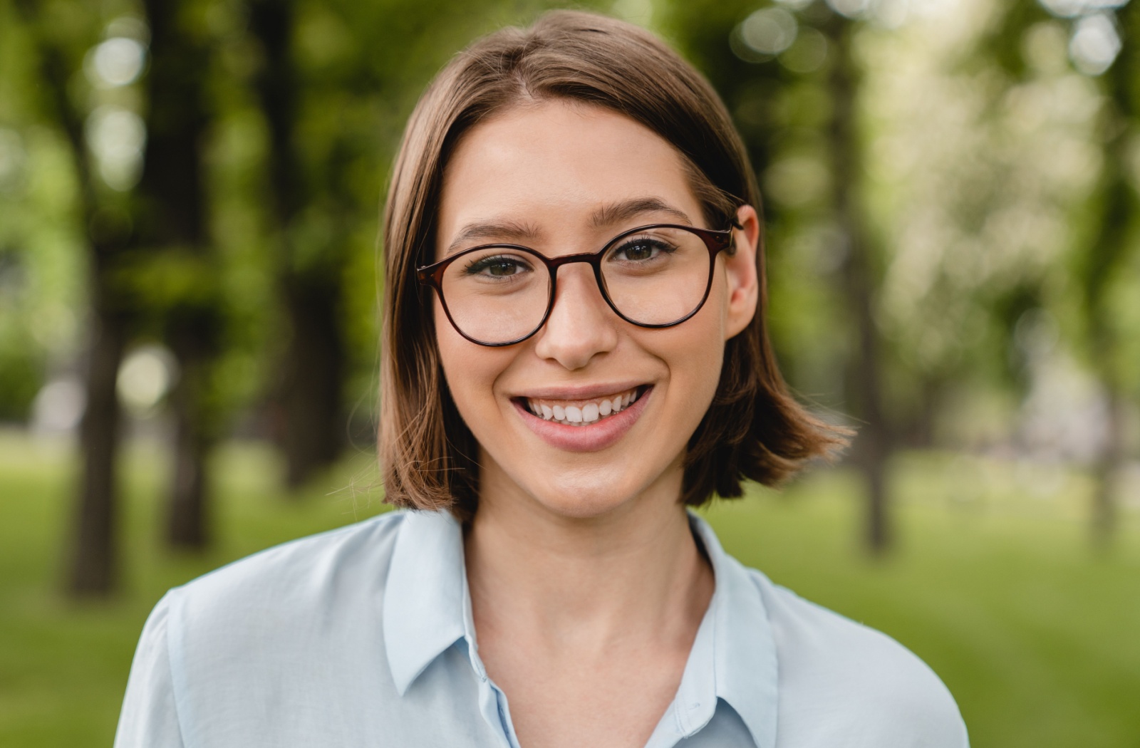 A person wearing glasses with Neurolens lenses smiles ahead while standing in a lush, green park.