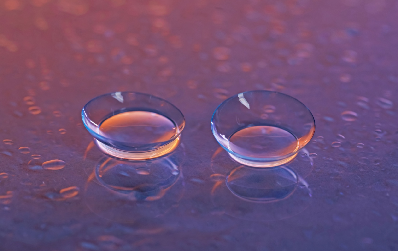 A pair of contact lenses sit on a purple background while moisturizing water droplets are scattered around them.
