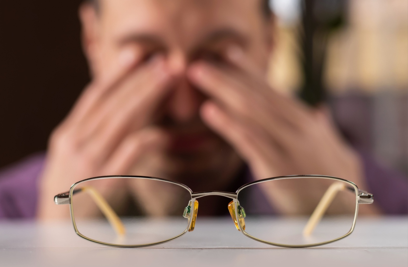 A pair of glasses rests in sharp focus on a surface, while a person in the blurred background rubs their eyes with both hands.