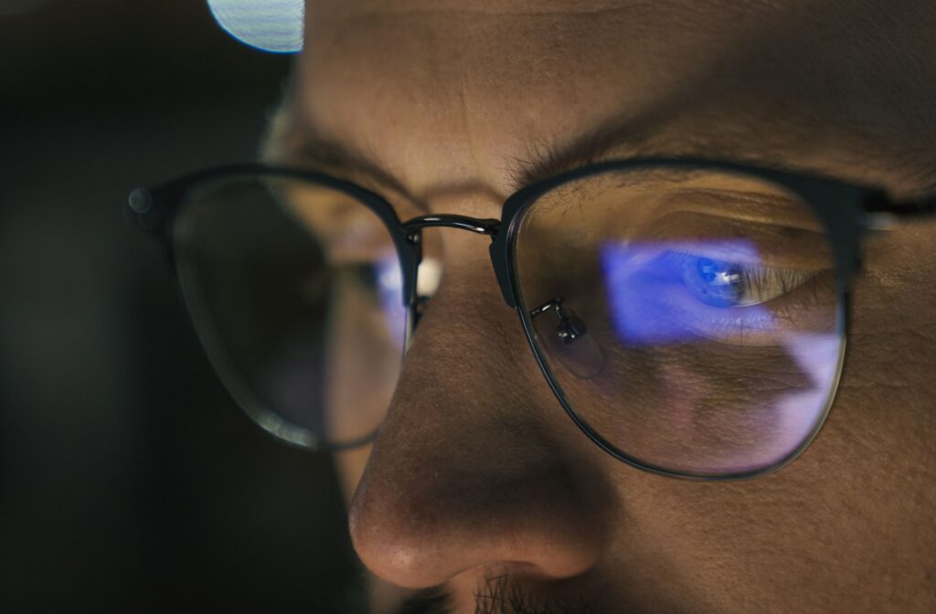 Close-up of a person’s eyes behind glasses, reflecting the blue glow of a screen in a dark room.