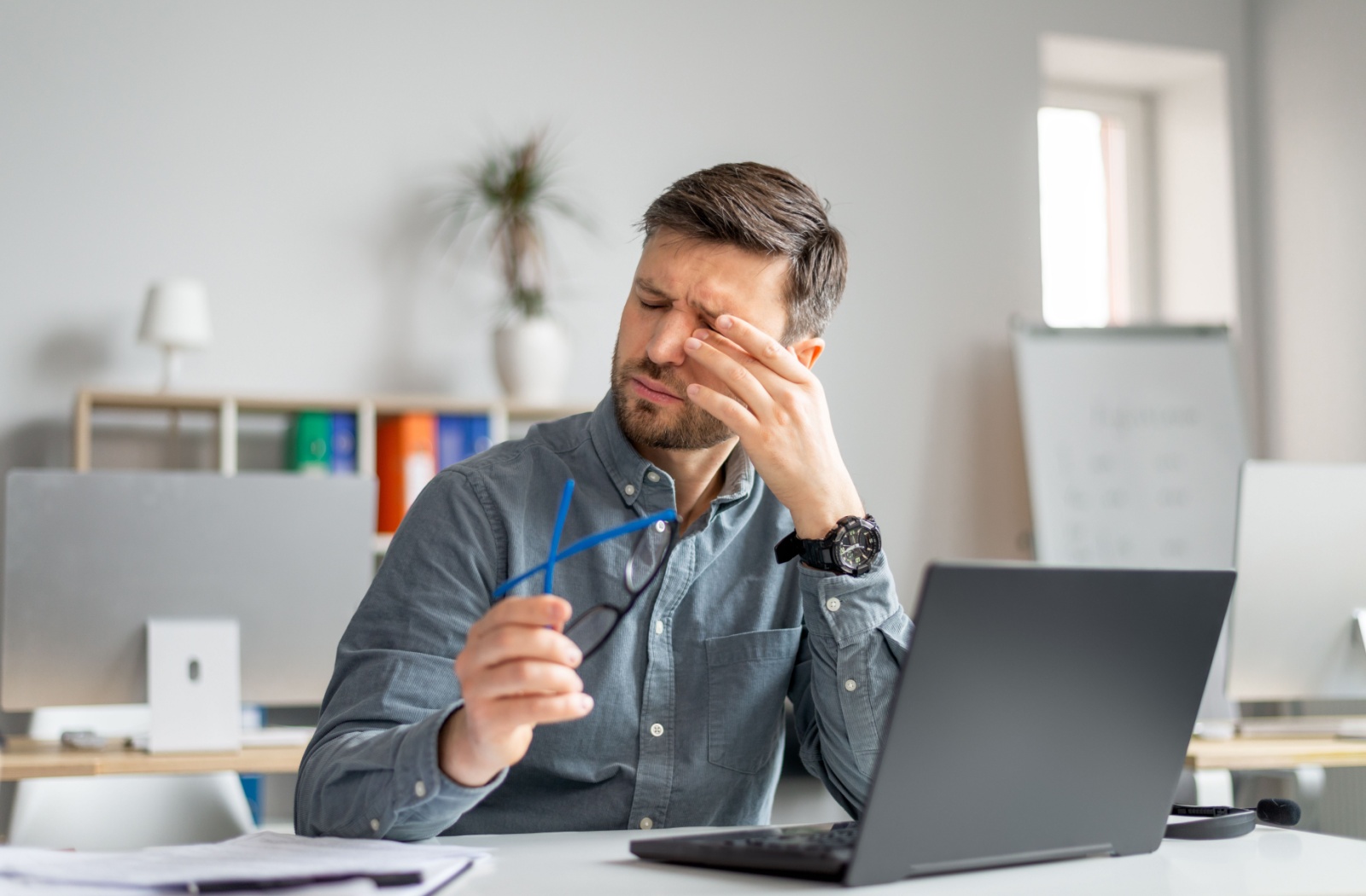 Man taking glasses off to rub his eyes in front of a computer while sitting at a desk
