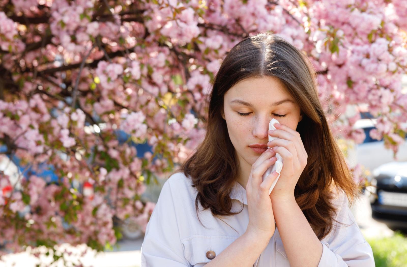 A person dabbing their eye with a tissue while standing in front of blossoming trees.